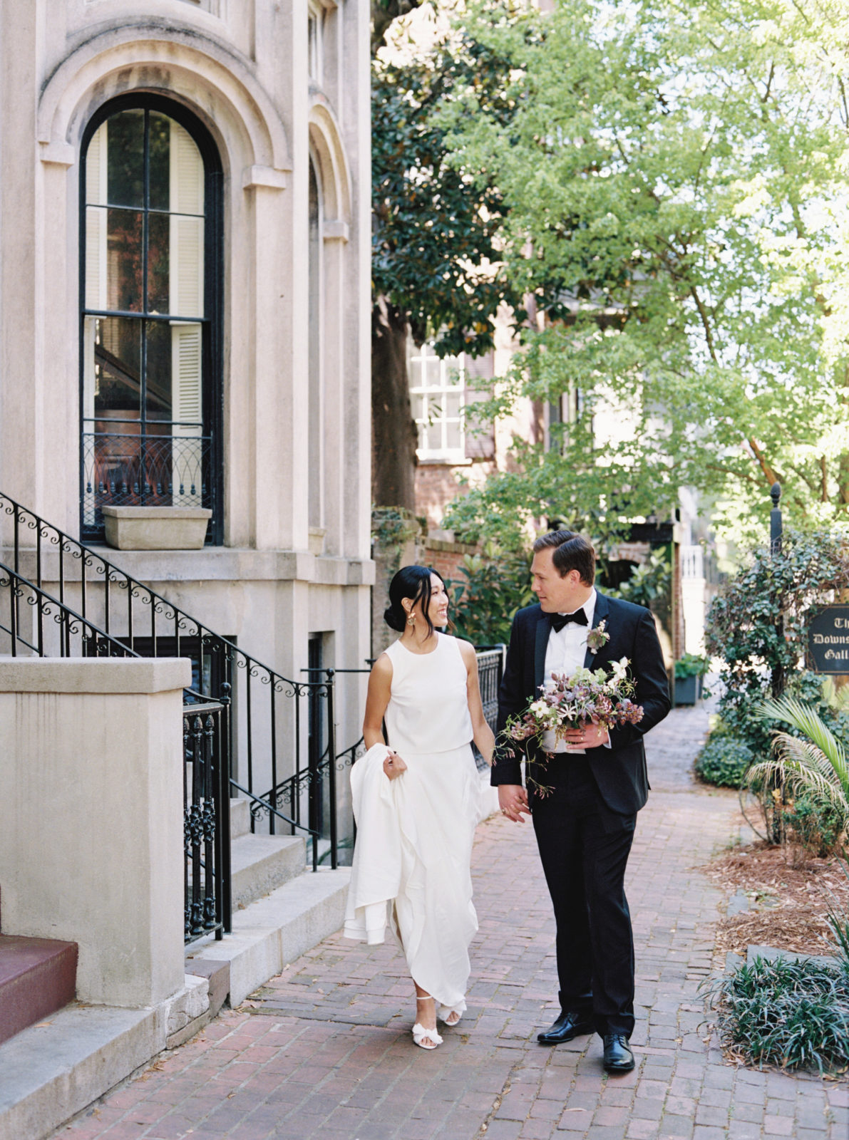 Couple goes for a walk in the streets of Savannah during their old world-inspired elopement. Brides hold train of her modern gown while groom holds bouquet.