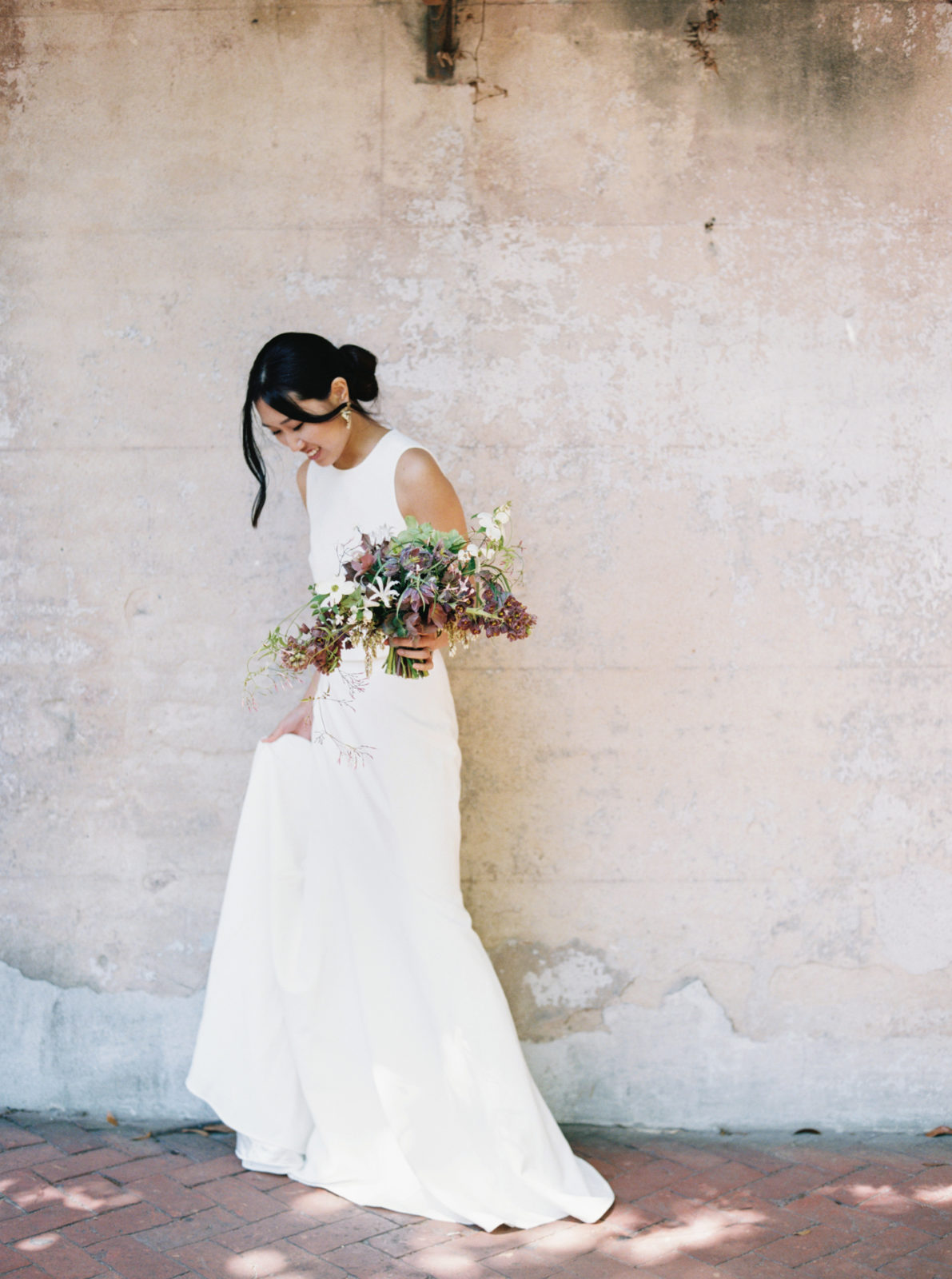 Bride holds dress and looks down while standing against rustic building holding a colorful bouquet in Savannah, Georgia.