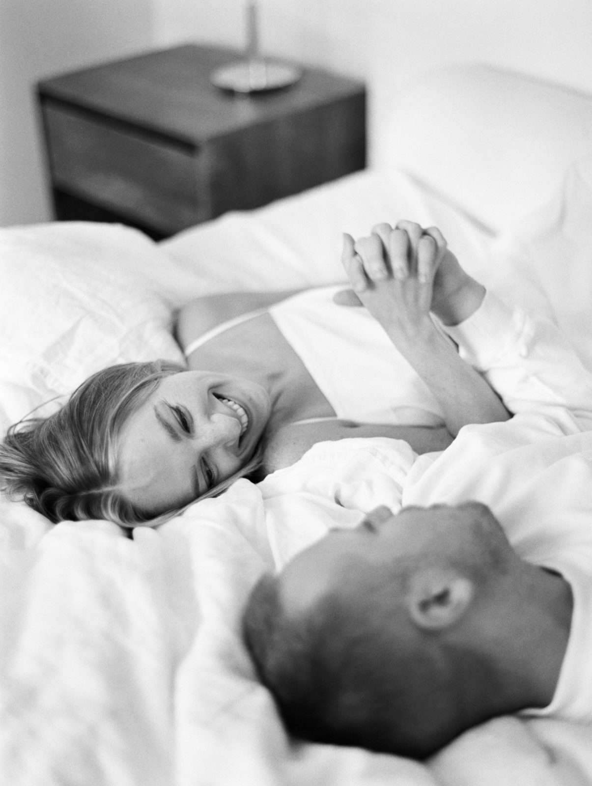 Wife smiles at husband while laying on bed holding hands. Black and white film during lifestyle in-home session.