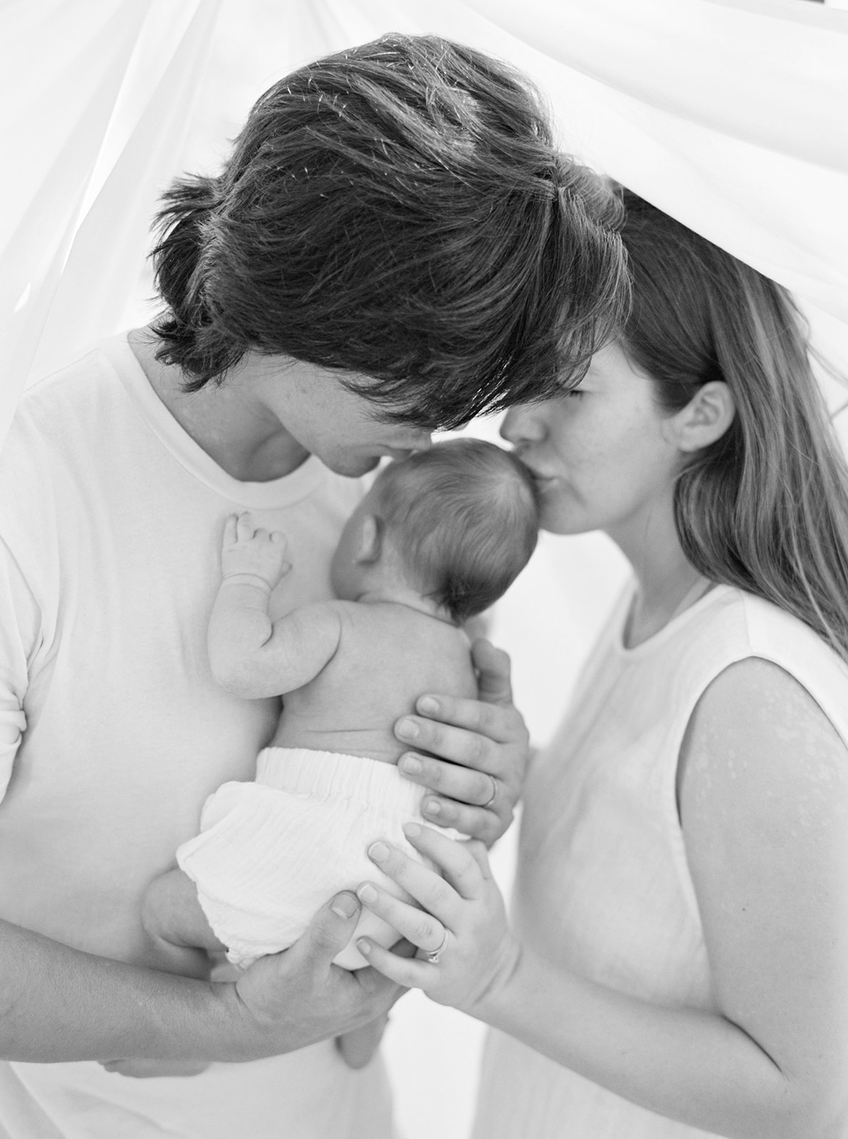 Black and white organic newborn portrait with mom and dad kissing newborn baby boy.