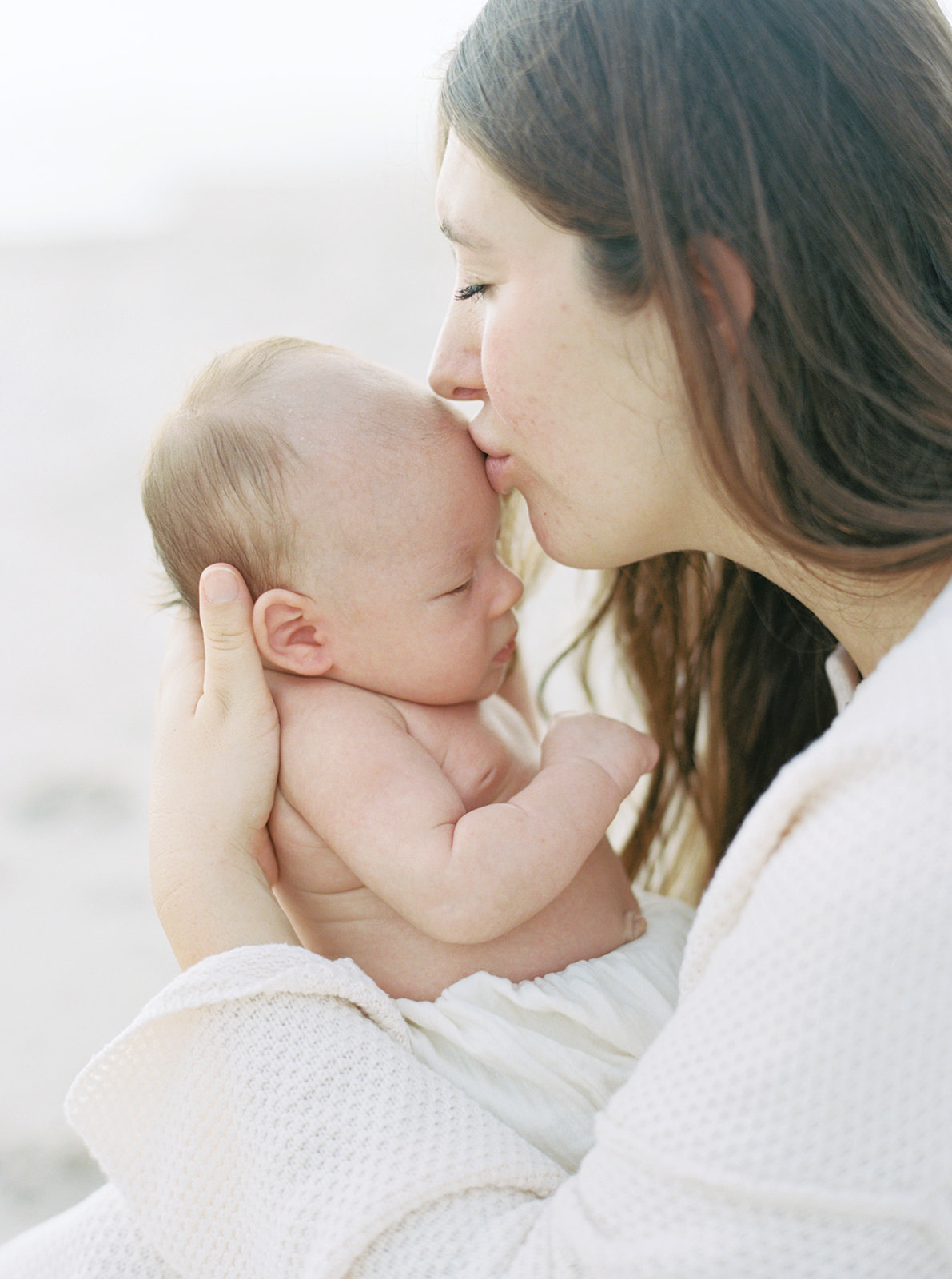 Mom gives baby a tender kiss on the forehead during organic newborn portraits on the beach.