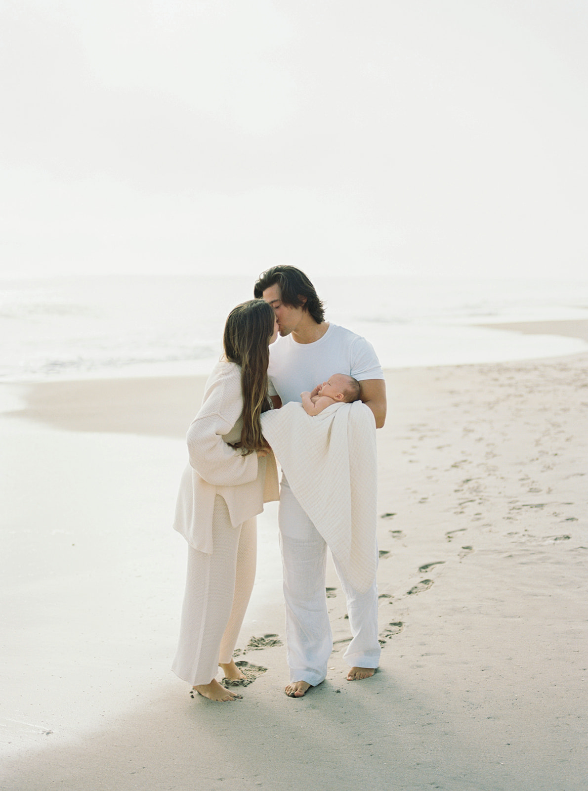 Mom and dad kissing on a hazy beach morning while dad is holding newborn baby wrapped in organic blanket.