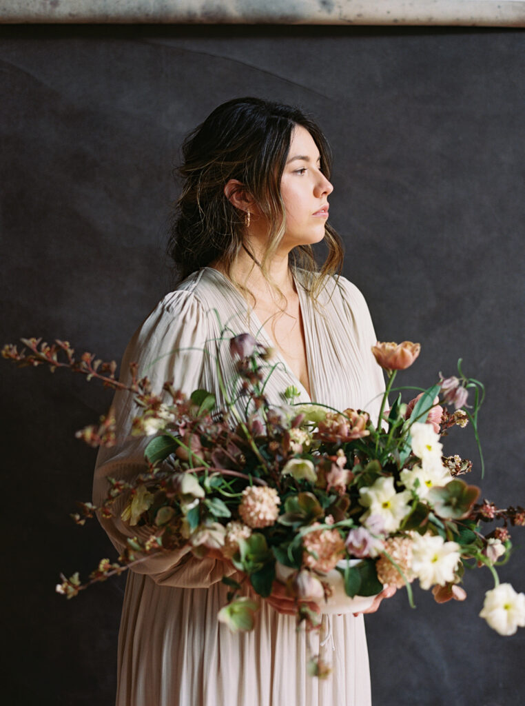 A portrait of a floral designer facing a window while holding an arrangement of moody toned flowers in front of a dark backdrop 