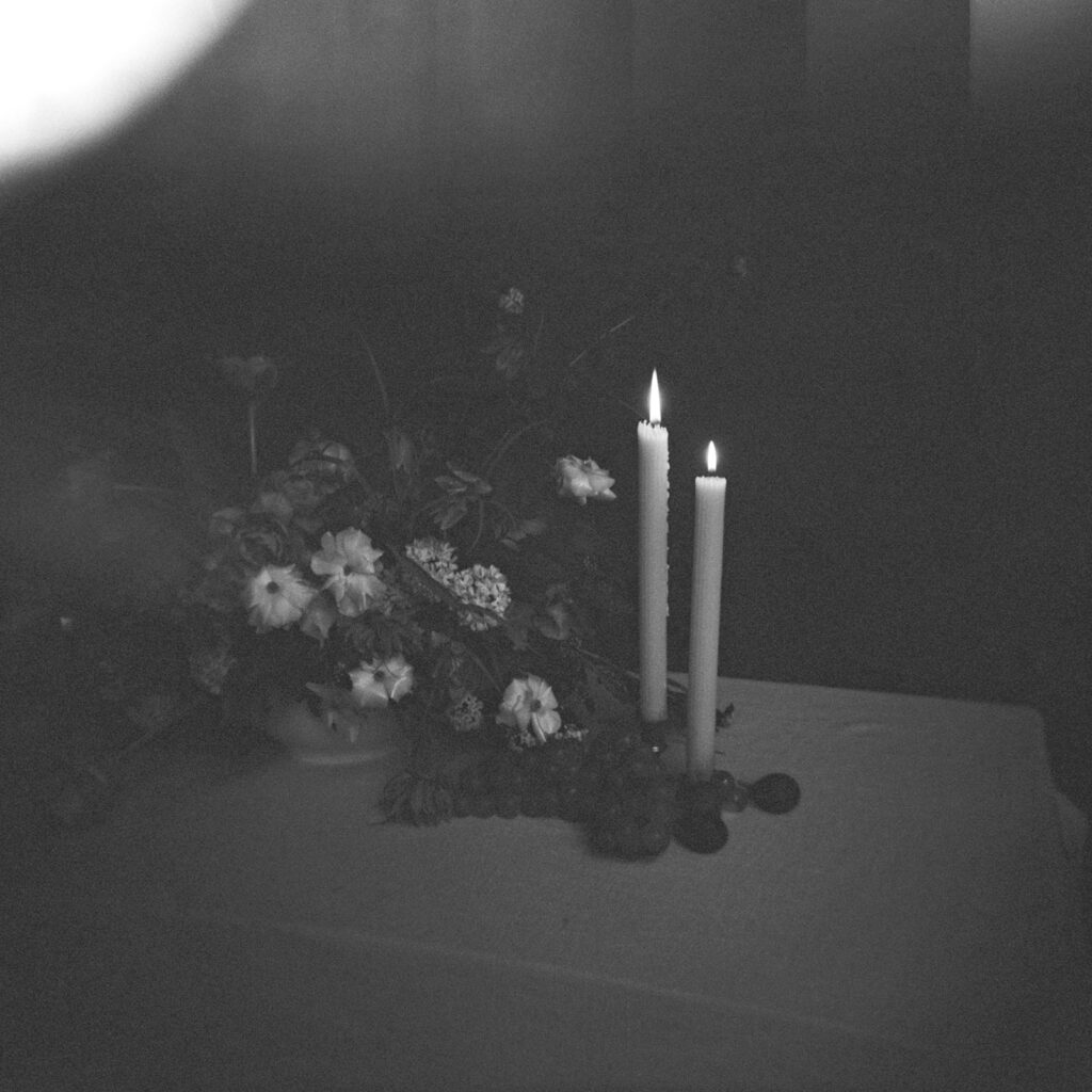A grainy black and white film photo of a candlelit table next to a floral arrangement