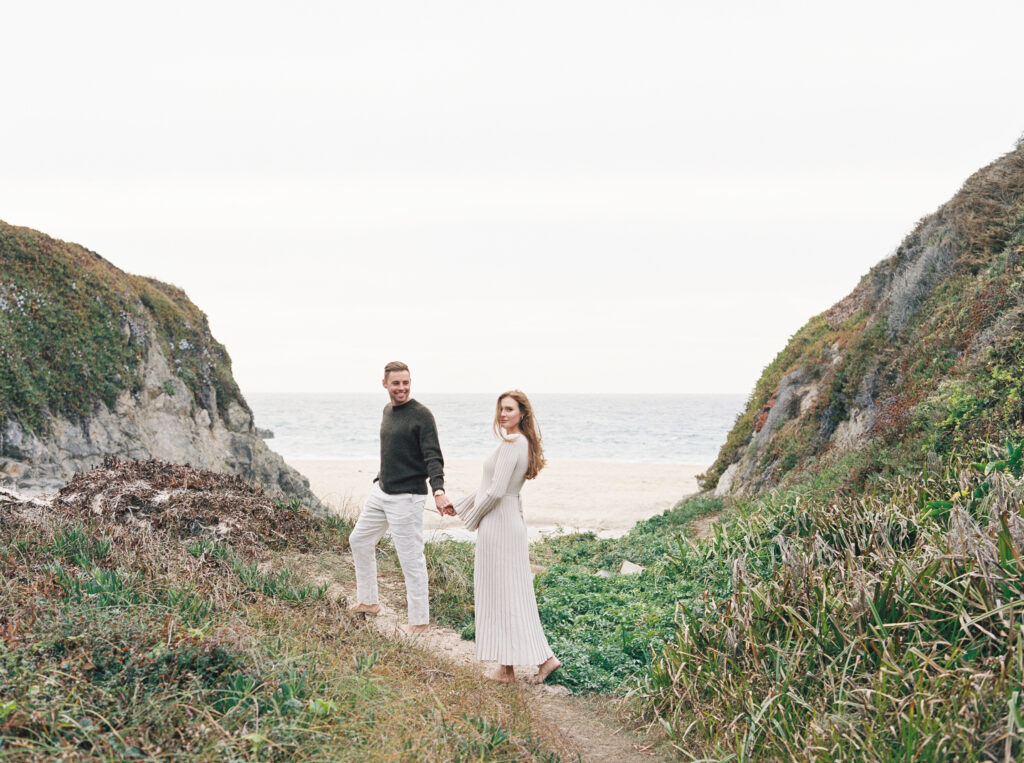 Couple walks with ocean in the background during their early maternity session in Big Sur at sunset