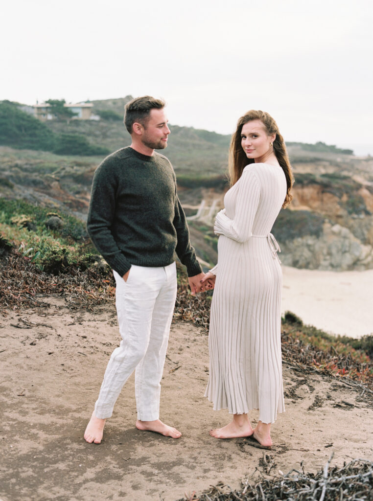 Couple stands on cliff overlooking Garrapata Beach during their early maternity session in Big Sur at sunset