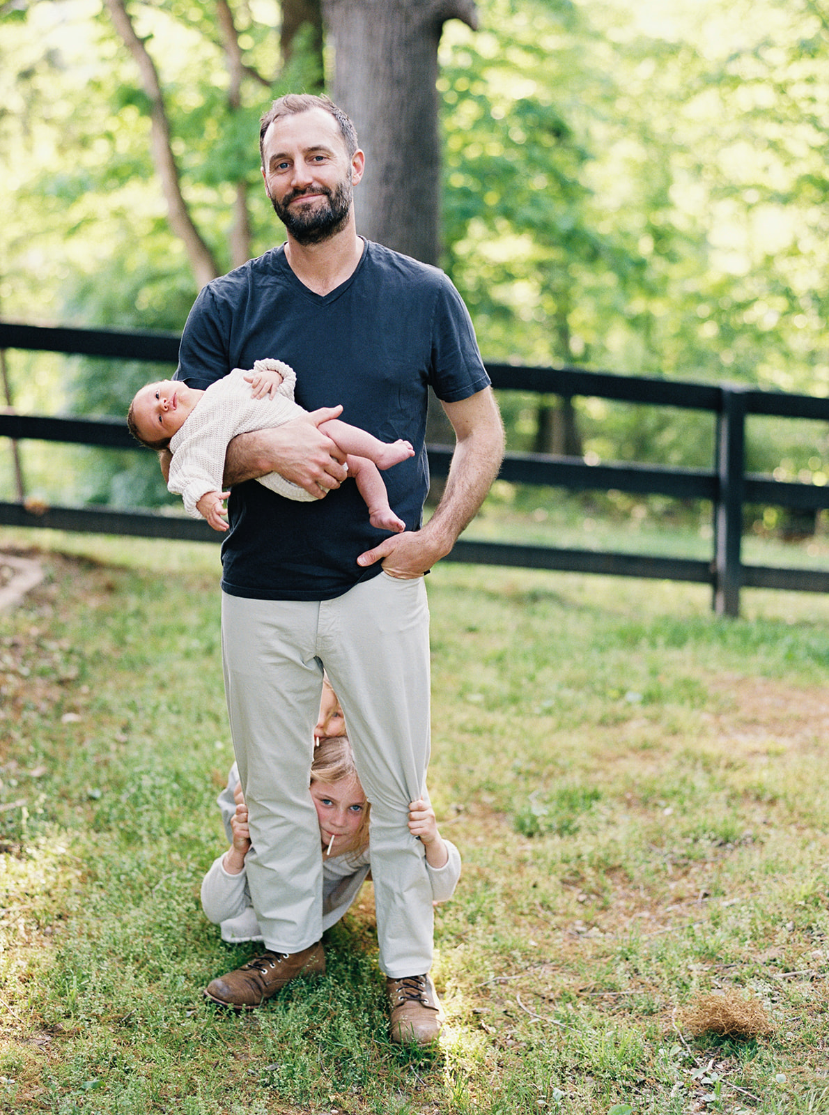 Father stands in the yard holding newborn son while daughters peek between his legs during their in-home family session in Nashville, Tennessee