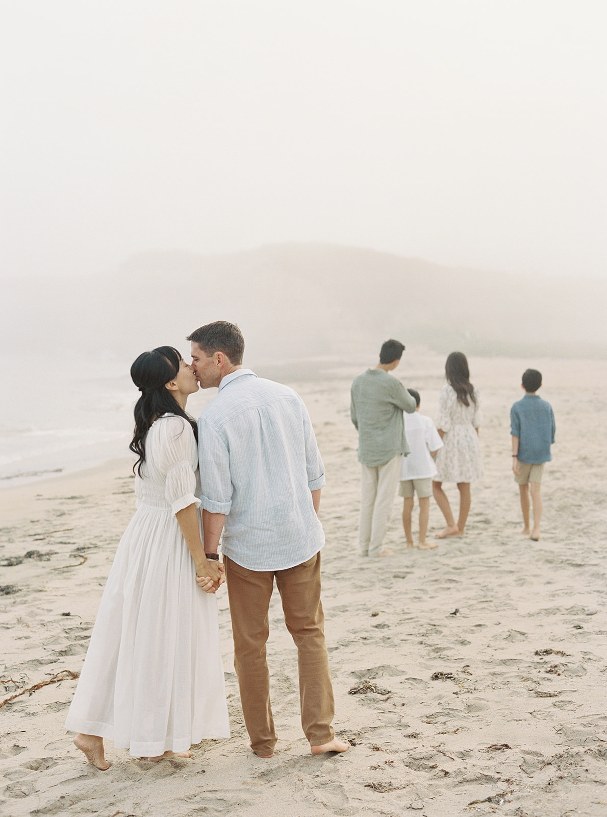 A Northern California family session featuring a mom and dad kissing while their kids walk ahead down the beach